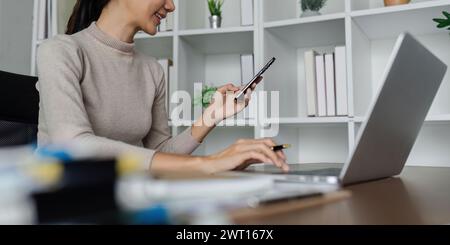 femme d'affaires utilisant un smartphone tout en travaillant sur ordinateur portable, synchroniser les données entre l'ordinateur et le gadget à la maison, utiliser les appareils d'entreprise et les affaires Banque D'Images