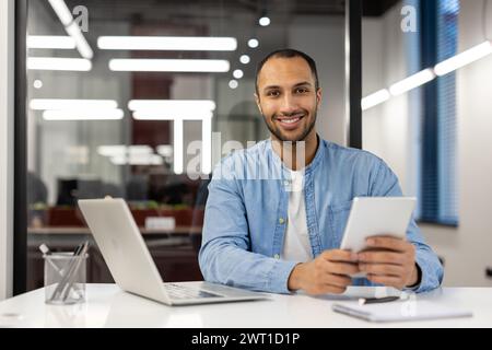Portrait d'un jeune homme afro-américain assis à un bureau dans un bureau moderne, tenant une tablette et souriant à la caméra. Banque D'Images