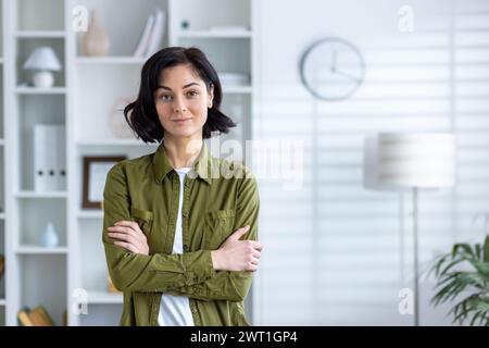 Portrait d'une jeune enseignante confiante debout à la maison dans une chemise verte, serrant ses mains sur sa poitrine et regardant la caméra. Banque D'Images
