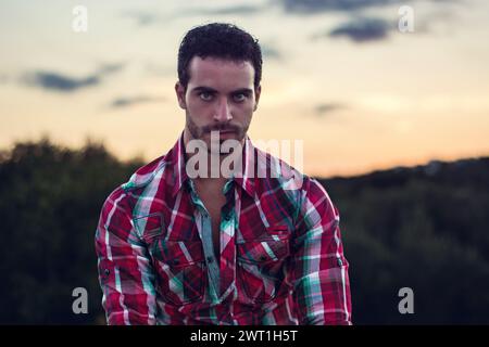 Un homme portant une chemise à carreaux rouge et vert se tient dehors. Il regarde droit devant lui, avec une expression détendue sur son visage, sur un fond simple. Banque D'Images