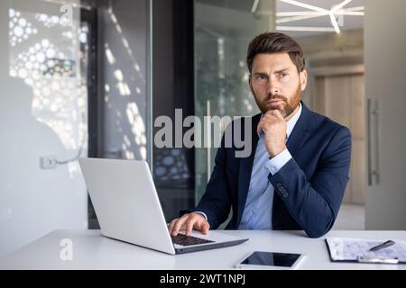 Portrait d'un jeune homme d'affaires sérieux assis dans un costume à un bureau dans un bureau moderne devant un ordinateur portable, regardant avec confiance sur le côté tout en tenant sa main près de son menton. Banque D'Images