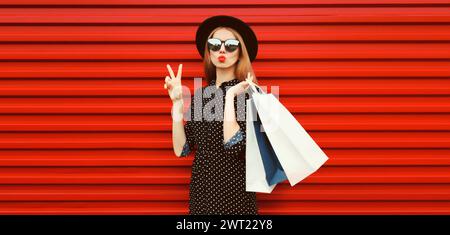 Portrait de belle jeune femme avec des sacs à provisions soufflant ses lèvres envoie un baiser d'air portant un chapeau rond noir sur fond rouge Banque D'Images