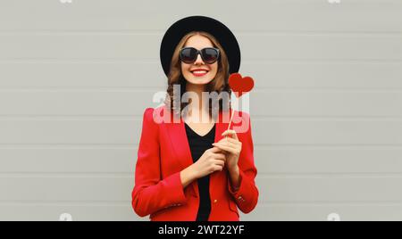 Belle femme heureuse avec sucette douce en forme de coeur rouge sur bâton dans un chapeau rond noir Banque D'Images