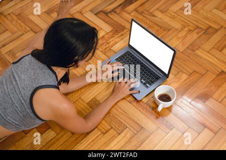 vue de dessus de la jeune femme brune méconnaissable sur le sol avec une tasse de café, couchée en écrivant à l'aide d'un ordinateur portable avec un espace de copie d'écran blanc Banque D'Images