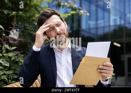 Photo en gros plan d'un jeune homme d'affaires choqué qui est dehors dans la rue, a reçu de mauvaises nouvelles par lettre, triste a fermé les yeux et se tient la tête. Banque D'Images