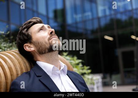 Photo en gros plan d'un jeune homme d'affaires pensif et fatigué assis à l'extérieur d'un immeuble de bureaux, appuyé sur un banc, levant les yeux, bouleversé et déçu. Banque D'Images