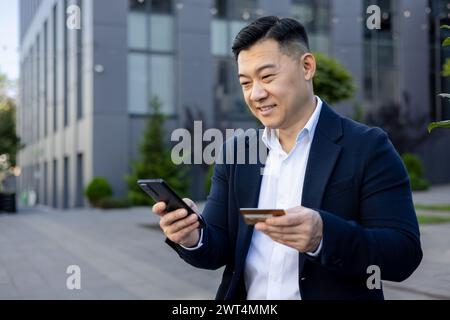 Dirigeant asiatique confiant qui gère les transactions financières avec une carte de crédit et un téléphone portable à l'extérieur des immeubles de bureaux modernes. Banque D'Images