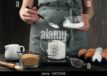 Femme ajoutant de la levure chimique dans la tasse à mesurer à la table en bois noir, gros plan Banque D'Images