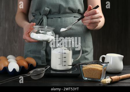 Femme ajoutant de la levure chimique dans la tasse à mesurer à la table en bois noir, gros plan Banque D'Images