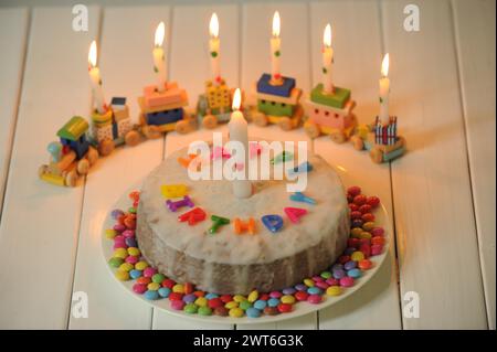 Ein Geburtstagskuchen mit brennenden Kerzen und dem Schriftzug Joyeux anniversaire umgeben von bunten Suessigkeiten, ein Geburtstagszug im Hintergrund Banque D'Images