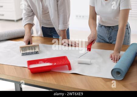 Femme et homme appliquant de la colle sur la feuille de papier peint à la table en bois à l'intérieur, gros plan Banque D'Images