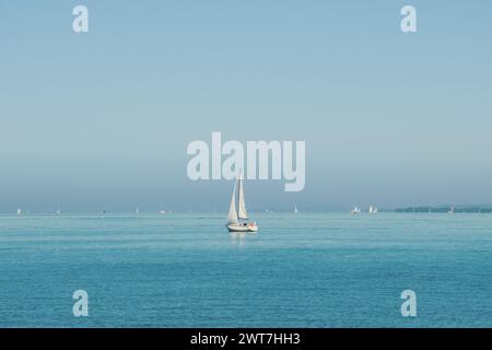 Petit voilier blanc dans le lac bleu avec beaucoup d'autres bateaux loin en arrière-plan. Yacht à voile avec un mât sur une journée ensoleillée dans le grand lac. Bleu eau bleu ciel bleu Banque D'Images