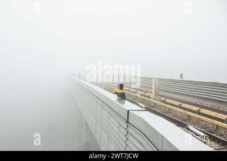 Ligne de métro surélevée se dégageant dans le brouillard. Rails sur un pont au-dessus d'une rivière - faible visibilité. Trajet matinal - attente d'un train. Vue en perspective Banque D'Images