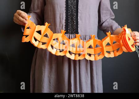 Femme en robe gothique grise tient une guirlande de papier. Guirlande de citrouille sur fond sombre. Décorations d'Halloween écologiques faites à la main. Banque D'Images