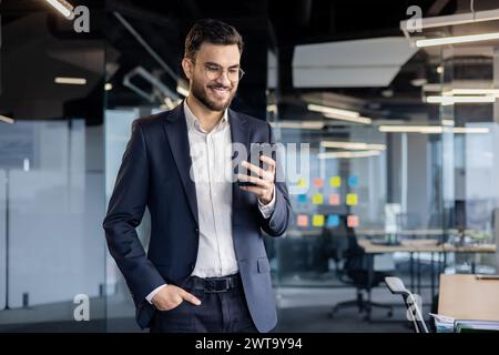 Un jeune homme d'affaires joyeux en costume interagit avec son smartphone dans un cadre de bureau lumineux et contemporain. Banque D'Images