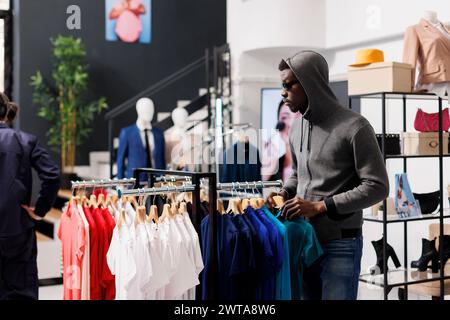 Homme afro-américain regardant de côté tout en volant la boutique moderne, vérifiant les vêtements à la mode. Voleur essayant de voler des marchandises élégantes, portant des lunettes de soleil et une capuche pour ne pas être reconnu Banque D'Images