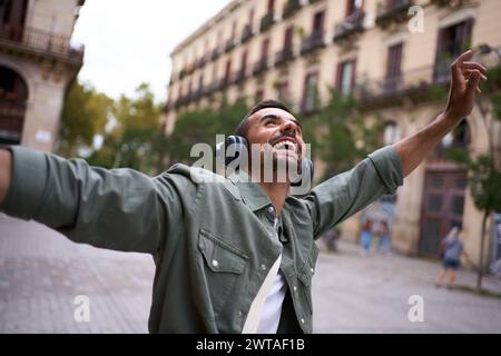 Jeune homme excité avec des écouteurs sur des stands dans la rue européenne de la ville de Barcelone avec les bras tendus Banque D'Images