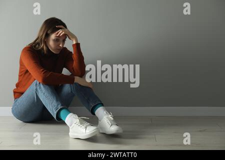 Triste jeune femme assise sur le sol près du mur gris à l'intérieur, espace pour le texte Banque D'Images