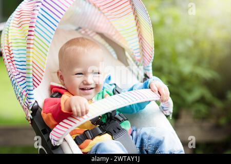 Bébé garçon dans un chandail blanc assis dans une poussette blanche sur une promenade dans un parc. Enfant en buggy arc-en-ciel colorée. Un petit enfant dans une poussette. Banque D'Images