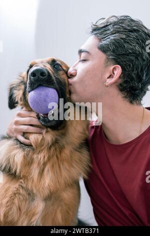 Un jeune homme latino montre de l'affection en embrassant son Berger allemand tenant une balle bleue Banque D'Images