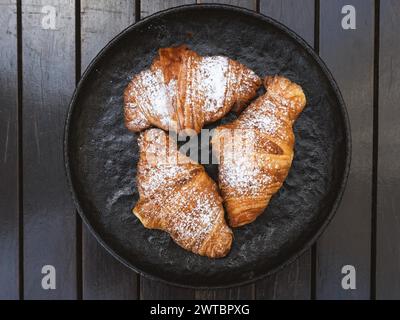 Trois croissants avec du sucre en poudre sur le dessus. Les croissants sont disposés dans un bol noir sur une table en bois Banque D'Images