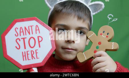 Mignon garçon avec pull de noël et chevreuil Alntlers tenant le signe Père Noël s'arrête ici et homme en pain d'épice. Isolé sur fond vert,. Photo de haute qualité Banque D'Images