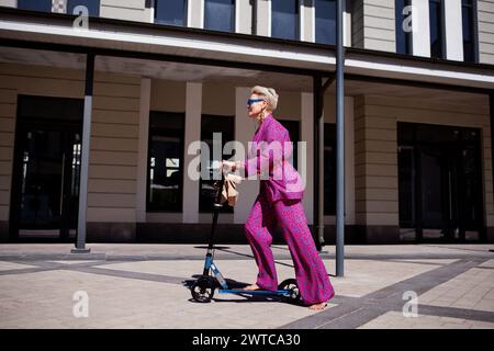 Élégamment habillée femme mature dans un pantalon rose vif et une veste roule autour de la ville sur un scooter, un frien actif, sain et éco Banque D'Images