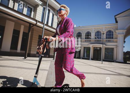 Élégamment habillée femme mature dans un pantalon rose vif et une veste roule autour de la ville sur un scooter, un frien actif, sain et éco Banque D'Images