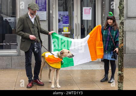 Leeds, Royaume-Uni. 17 MARS 2023. Chien drapé de drapeau irlandais pose aux côtés de propriétaires tenant le drapeau irlandais alors que la parade de Leeds de St Patricks se déplace dans le centre-ville. Crédit Milo Chandler/Alamy Live News Banque D'Images