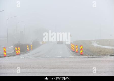 une voiture solitaire roule sur une route déserte et brumeuse flanquée de panneaux de signalisation, avec un environnement à peine visible en raison de l'épais brouillard Banque D'Images