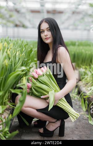 Jeune femme debout dans une grande serre et tenant des tulipes en fleurs dans ses mains. Banque D'Images