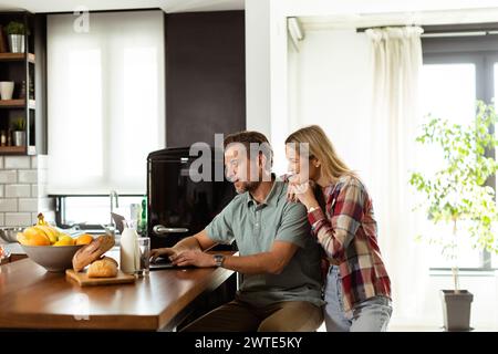 Un couple joyeux profite d'un moment léger dans leur cuisine ensoleillée, travaillant sur ordinateur portable entouré d'un petit déjeuner sain Banque D'Images
