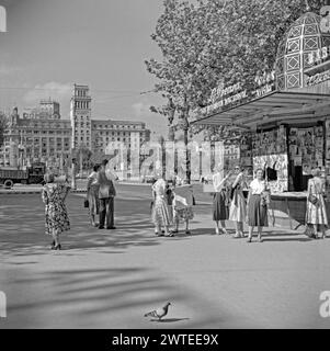 Une vue des touristes devant un kiosque à journaux à l'extrémité nord de Las Ramblas donnant sur la Plaça de Catalunya, Barcelone, Espagne en 1955. Plaça de Catalunya (place de Catalogne ou Plaza de Cataluña) est une grande place du centre de Barcelone qui est généralement considérée comme son centre-ville. La place occupe une superficie d'environ 50 000 mètres carrés. Il est surtout connu pour ses fontaines et statues et les troupeaux de pigeons qui se rassemblent au centre – une photographie vintage des années 1950. Banque D'Images