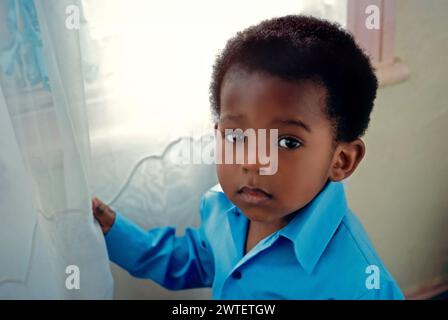 Bébé de 4 ans noir afro-américain garçon par fenêtre à la maison semble solennel et pensif, mais très mignon beau et attrayant, regardant la caméra, éclairé seulement par la lumière naturelle de la fenêtre. Banque D'Images