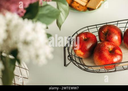 Un panier métallique rempli de pommes fraîches se trouve à côté d'un vase rempli de fleurs colorées, créant un affichage vibrant et invitant. Banque D'Images