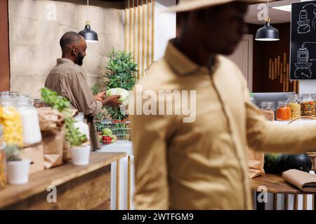 Un jeune homme noir remettant un chou au commerçant à la caisse. Des aliments frais et sains dans un emballage durable. Concentrez-vous sur l'arrière-plan. Banque D'Images