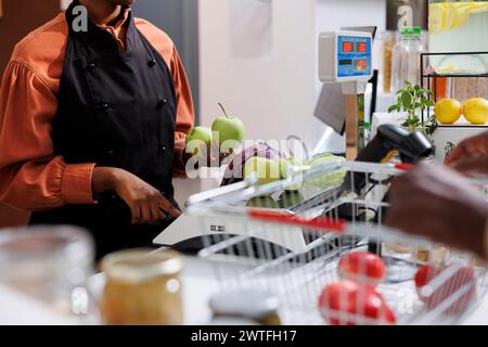 Accent sélectif sur la vendeuse afro-américaine tenant des pommes vertes et pesant un chou sur une balance. Les clients peuvent acheter une variété d'articles sains, y compris des fruits et des légumes. Banque D'Images