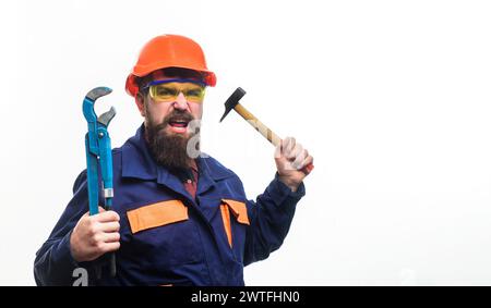 Constructeur en colère dans le casque avec clé à tube réglable et marteau. Homme barbu dans la construction uniforme et casque de protection avec clé réglable Banque D'Images