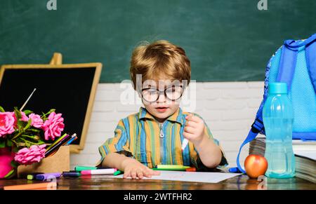 Mignon élève d'école primaire en classe. Petit écolier dessine au bureau. Fournitures scolaires et papeterie. Petit garçon étudiant dans des lunettes peignant avec Banque D'Images