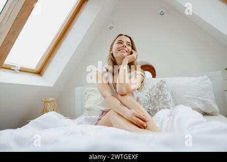 Une femme avec le sourire est assise sur un lit sous une lucarne, son bras reposant sur sa jambe Banque D'Images