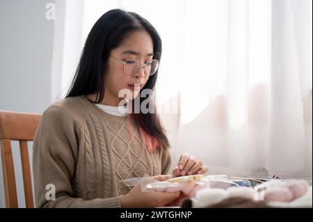 Une jeune femme asiatique portant des lunettes se concentre sur l'enfilage d'un motif sur un cadre de broderie, la couture à la main sur le tissu, tout en étant assis à une table par th Banque D'Images