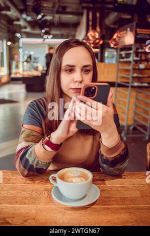 Une femme navigue attentivement son téléphone, une tasse de café à proximité, dans l'ambiance accueillante d'un café local Banque D'Images