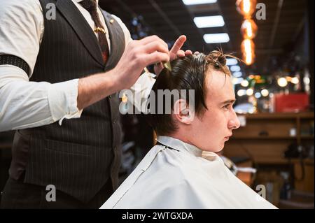 Barbier donnant la coupe de cheveux à un jeune homme dans un salon de coiffure. Barbier professionnel bien habillé en gilet et chemise blanche avec manches retroussées, se concentrant sur la coupe des cheveux, à l'aide d'un peigne et de ciseaux. Banque D'Images