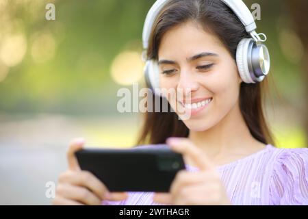 Femme heureuse regardant la vidéo sur le téléphone portant casque dans un parc Banque D'Images