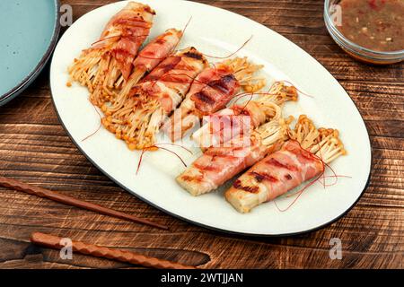 Champignon Enoki ou enokitake rôti au bacon et à la sauce sur une table en bois. Banque D'Images