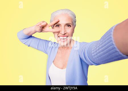 Une dame âgée fait un selfie sur l'appareil photo d'un smartphone faisant un v-sign près des yeux isolés sur fond violet clair avec un espace de copie pour le texte Banque D'Images