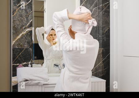 Femme en peignoir enveloppant les cheveux mouillés avec une serviette devant un miroir Banque D'Images