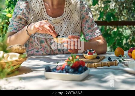 Femme anonyme faisant des mini-sandwichs sucrés pour un petit déjeuner sain dans le jardin. Toast avec fromage à la crème, myrtille, fraise, flocons de noix de coco et h. Banque D'Images