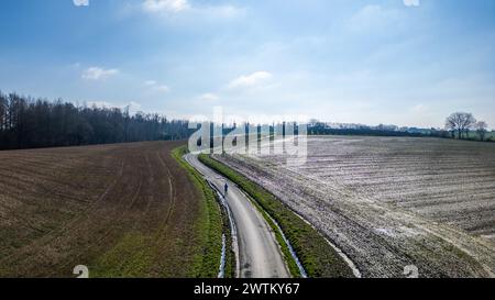 L'image présente une vue aérienne d'une route sinueuse et étroite serpentant à travers de vastes champs. Les champs semblent dormants avec des restes de neige ou de gel, suggérant que cette photo a été prise pendant les mois les plus froids. Une ligne d'arbres peut être vue au loin, marquant l'horizon sous un ciel bleu presque clair avec un minimum de nuages. La route sert de ligne directrice, attirant le regard à travers la composition, et il n'y a pas de véhicules ou de personnes visibles, donnant un sentiment de solitude et de tranquillité. Perspective aérienne de la route courbe au milieu des champs. Photo de haute qualité Banque D'Images