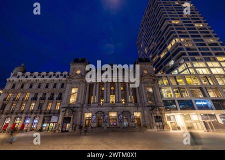 Anvers, Belgique, le 25 janvier 2024, cette image capture le contraste saisissant entre l'architecture historique de l'Opéra d'Anvers et la tour moderne qui l'entoure. Le bâtiment de l'Opéra, éclairé par de douces lumières, arbore un air de grandeur historique, tandis que le bâtiment contemporain adjacent présente des façades en verre qui reflètent le ciel du soir. La vie urbaine animée est visible à travers le mouvement des gens sur la place, avec le ciel bleu de l'heure ajoutant une toile de fond tranquille à cette scène urbaine. Crépuscule vue sur l'Opéra d'Anvers et l'édifice moderne de grande hauteur. Q élevé Banque D'Images